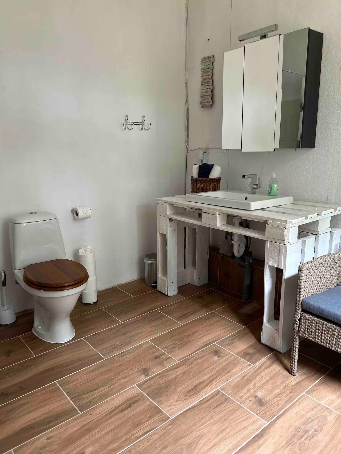 Bad mit WC, Waschtisch aus Palettenholz mit weißem Aufsatzbecken und Spiegelschrank; Boden mit Fliesen in Holzoptik, rechts ein Korbstuhl.