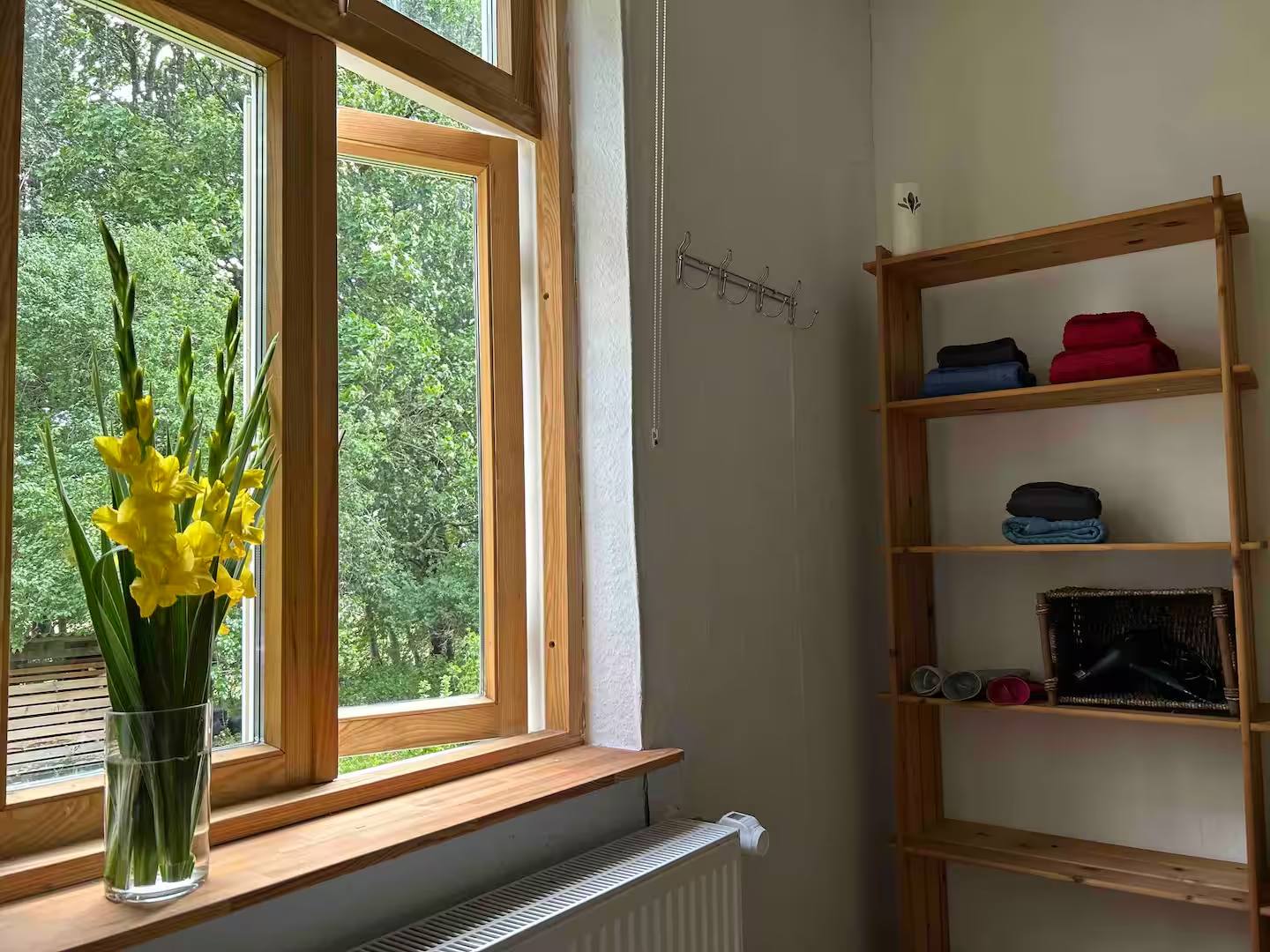 Geöffnetes Holzfenster mit Blick auf Bäume, auf der Fensterbank eine Vase mit gelben Gladiolen; rechts ein Regal und eine Garderobenleiste.