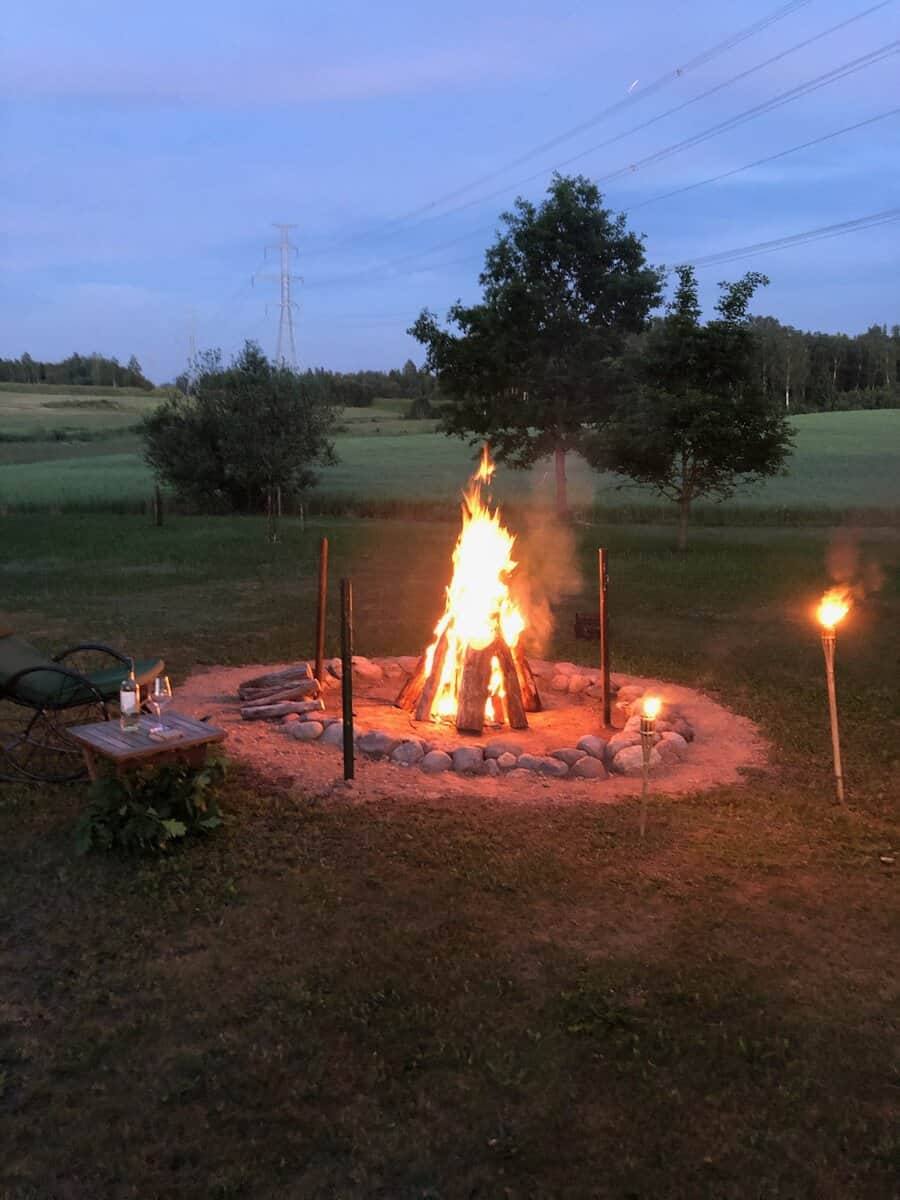 Brennende Feuerstelle mit zwei Gartenfackeln in der Dämmerung, dahinter Baumsilhouetten und offene Wiese.