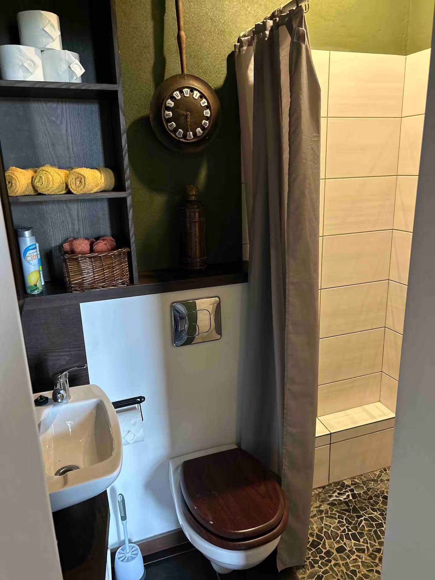 Gäste-WC mit Handwaschbecken, WC und gefliester Dusche hinter einem Vorhang; links ein olivgrünes Regal mit gerollten Handtüchern.