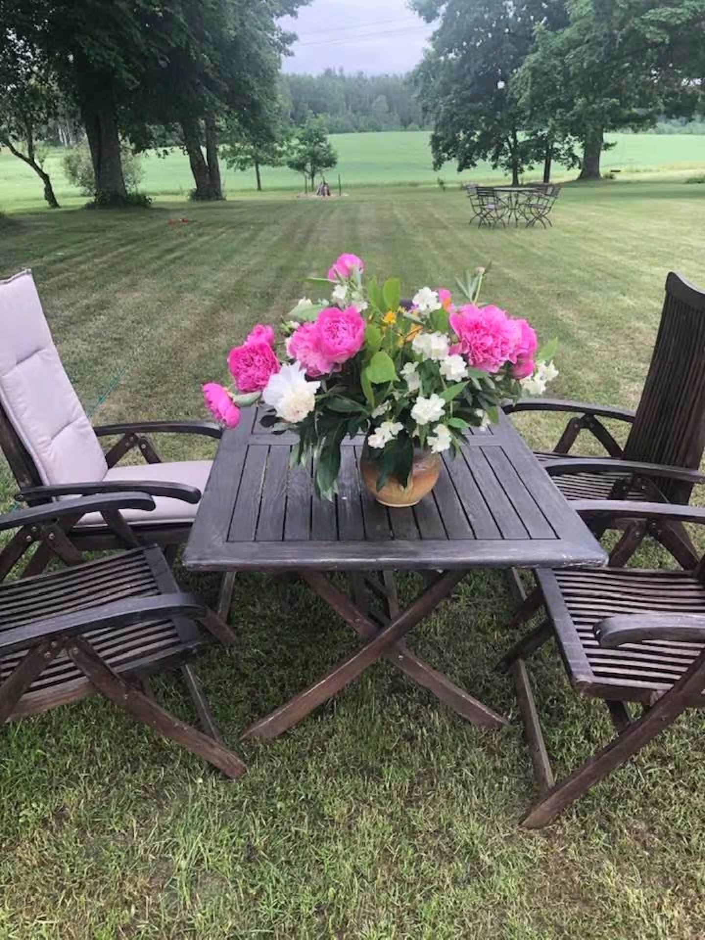 Gartentisch mit einem Strauß rosa und weißer Pfingstrosen, links und rechts Klappstühle, dahinter die weite Rasenfläche.