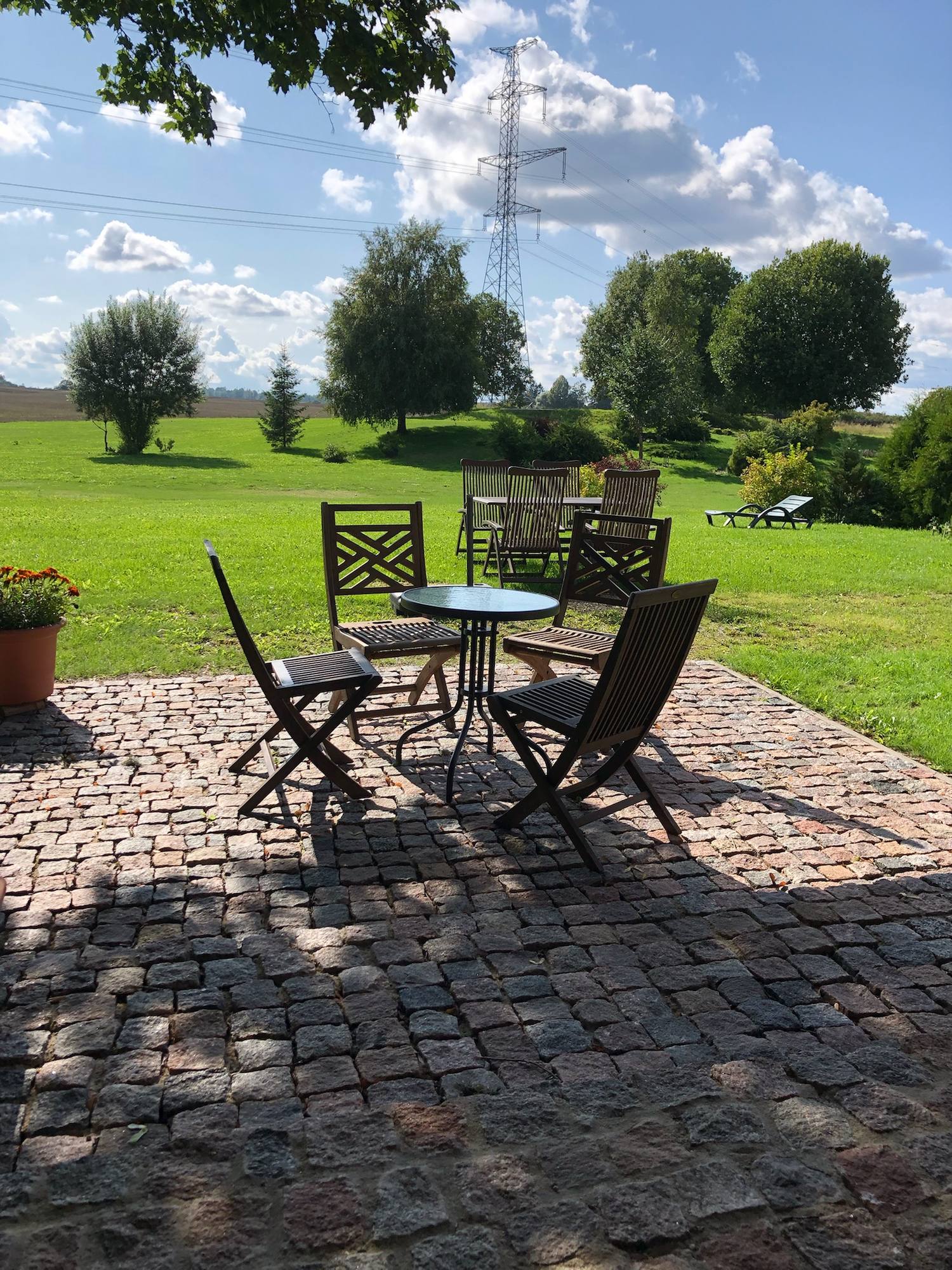 Gepflasterte Terrasse aus Natursteinen mit rundem Gartentisch und drei Klappstühlen, dahinter Rasen und eine Baumreihe.