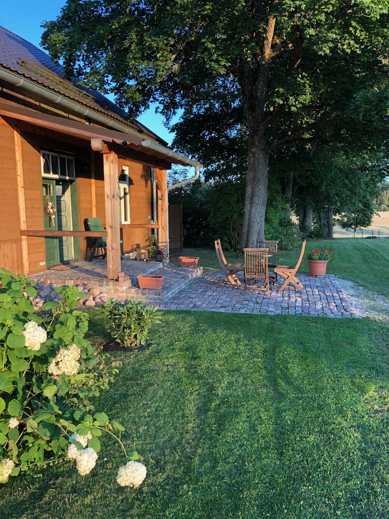 Seitlicher Blick auf Haus und gepflasterten Vorplatz, im Vordergrund blühende weiße Hortensien.