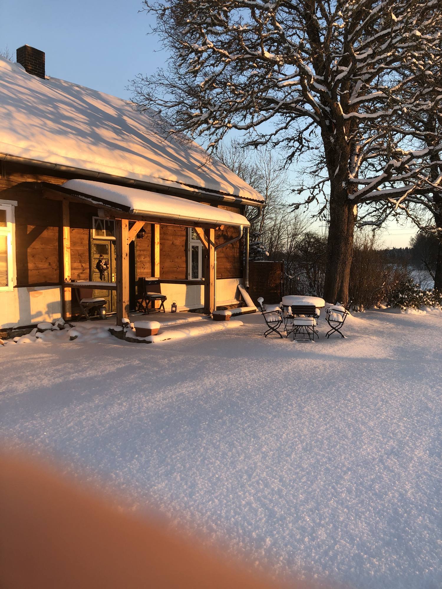 Das Haus im Schnee bei Abendlicht, die Fenster hinter dem Vordach sind erleuchtet; davor eine schneebedeckte Terrasse und eine kahle Eiche.