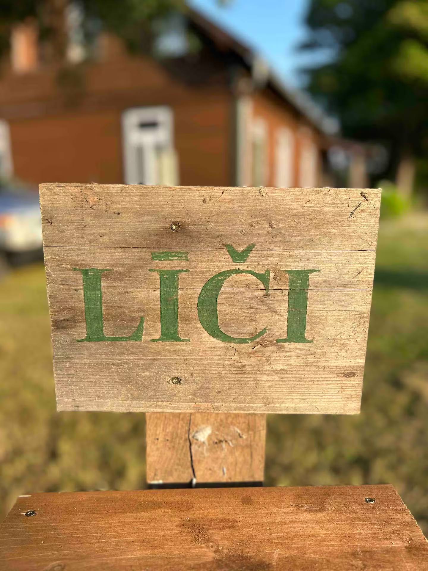 Holzschild mit der eingefrästen Aufschrift Līči auf einem Pfosten im Gras; im unscharfen Hintergrund das Haus.