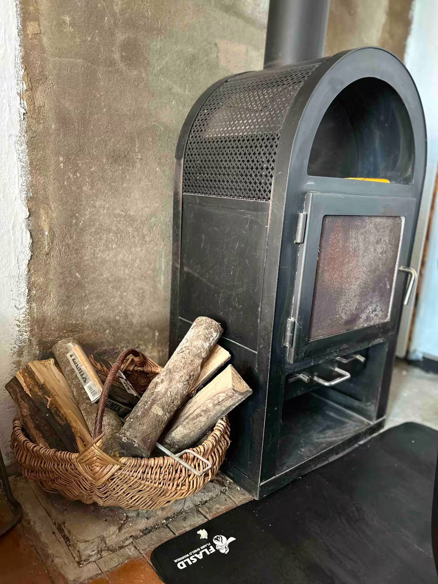 Freistehender schwarzer Holzofen mit Sichtscheibe an einer verputzten Wand, daneben ein Korb mit gespaltenem Feuerholz.