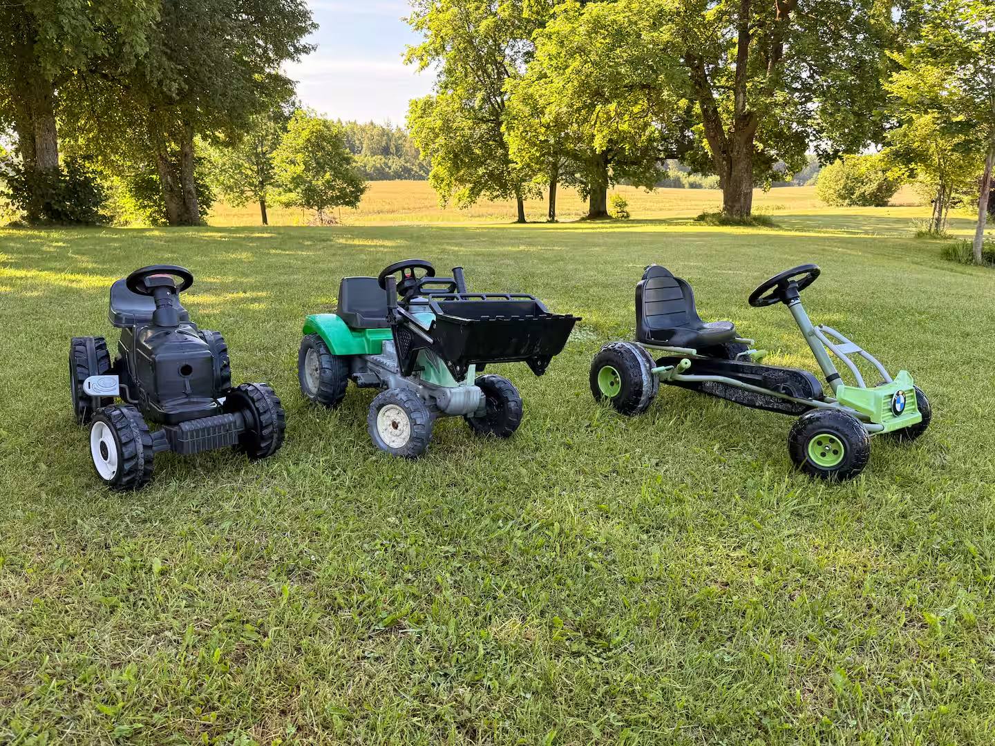 Drei Aufsitzfahrzeuge für Kinder, darunter ein Tretlader und ein Gokart, auf dem Rasen unter Bäumen.