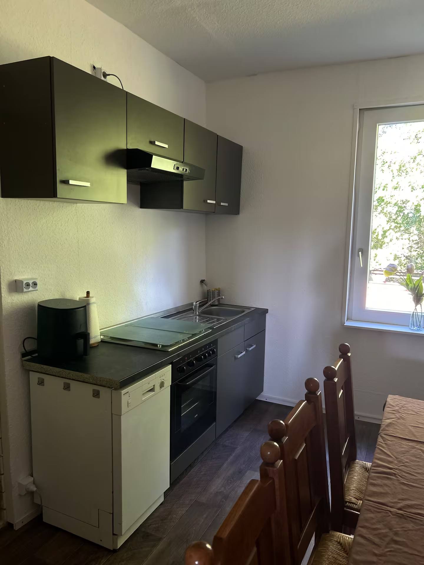 Küchenzeile mit Kochfeld, Einbaubackofen, Spüle und Arbeitsplatte, darüber Oberschränke mit Dunstabzugshaube; rechts ein Fenster.