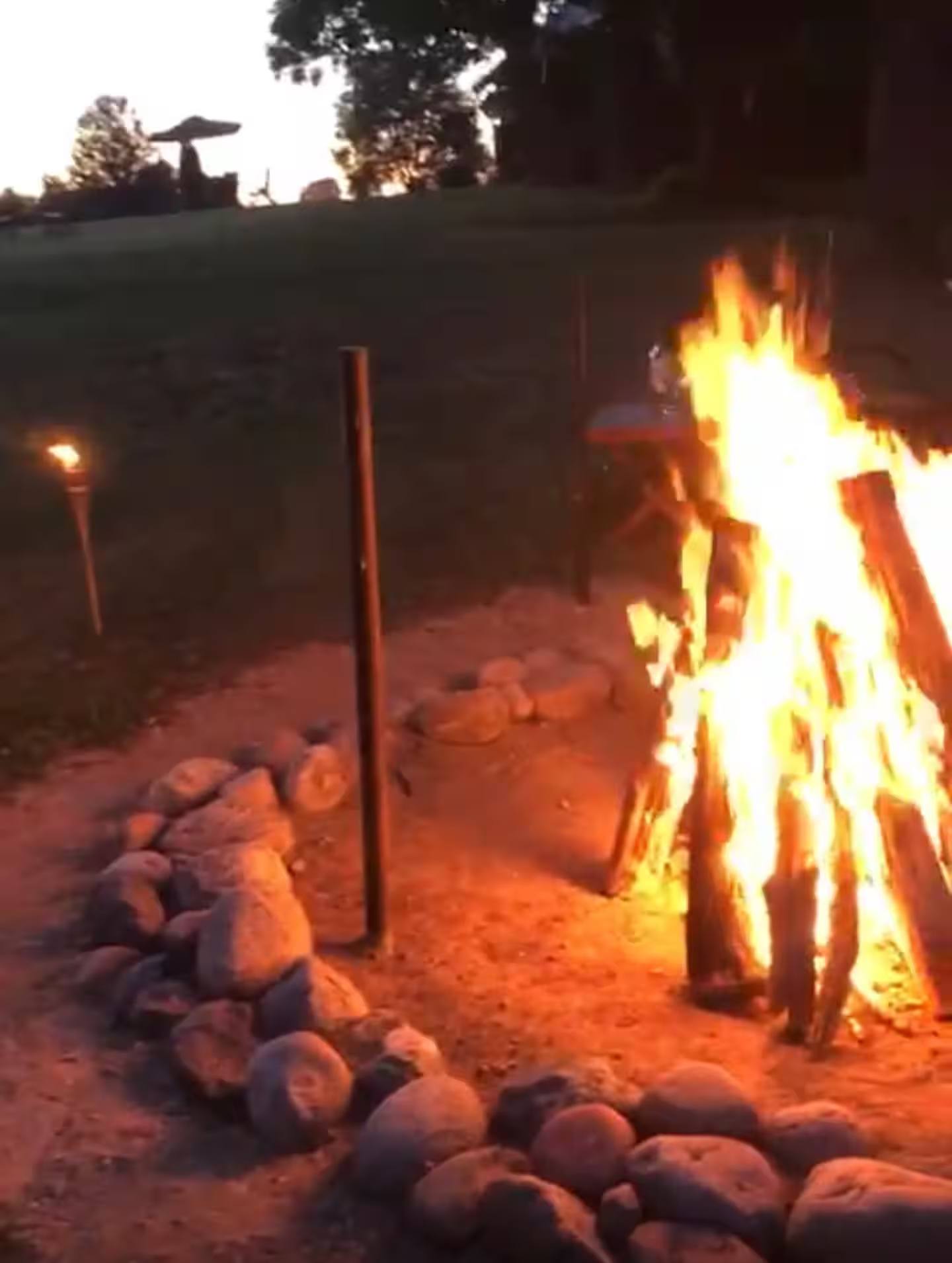Hohes offenes Feuer in einer mit Feldsteinen gefassten Feuerstelle, daneben eine brennende Gartenfackel.