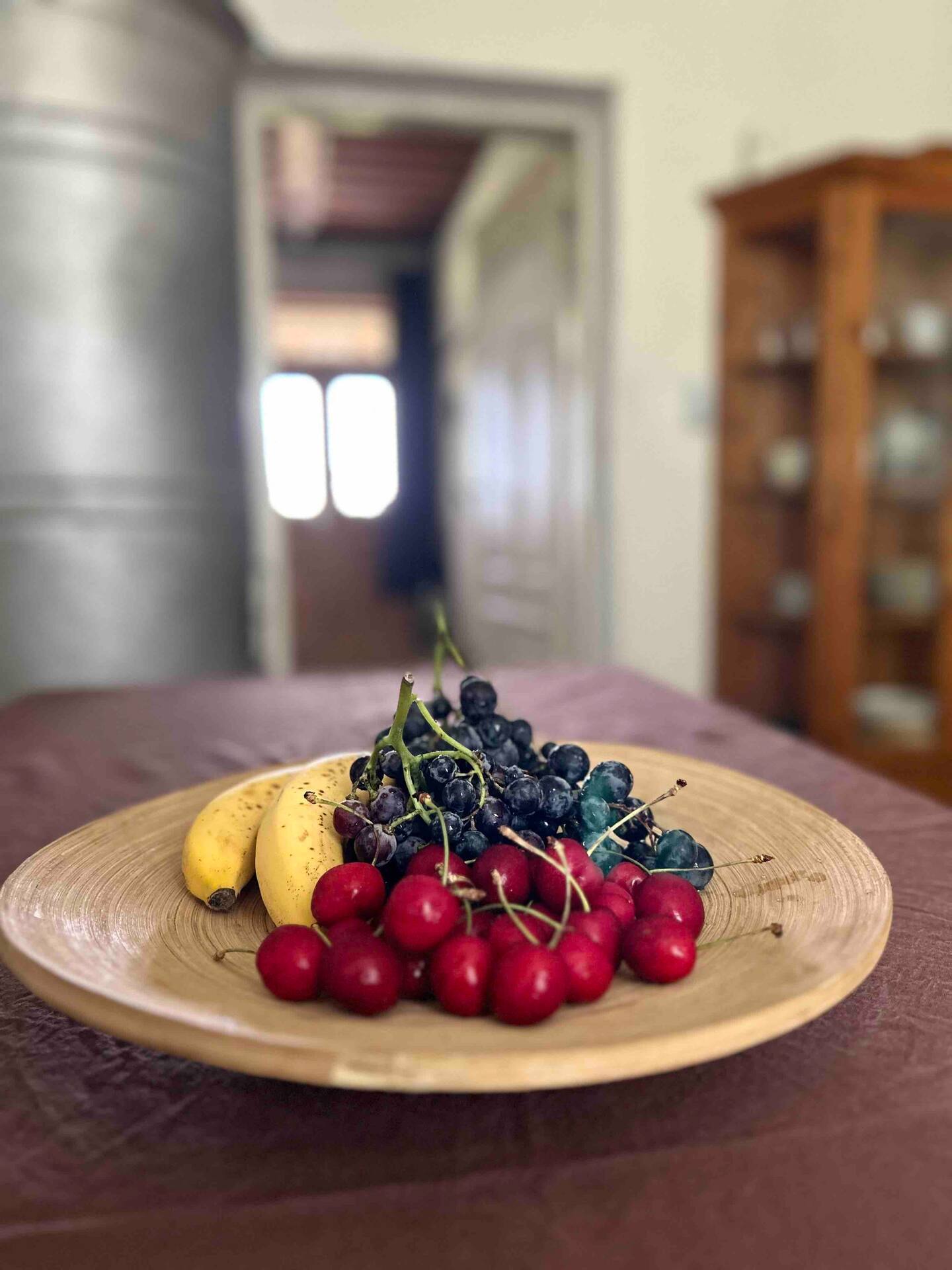 Holzschale mit Bananen, Kirschen und Weintrauben auf einem Esstisch, im unscharfen Hintergrund ein Durchgang und ein Buffetschrank.