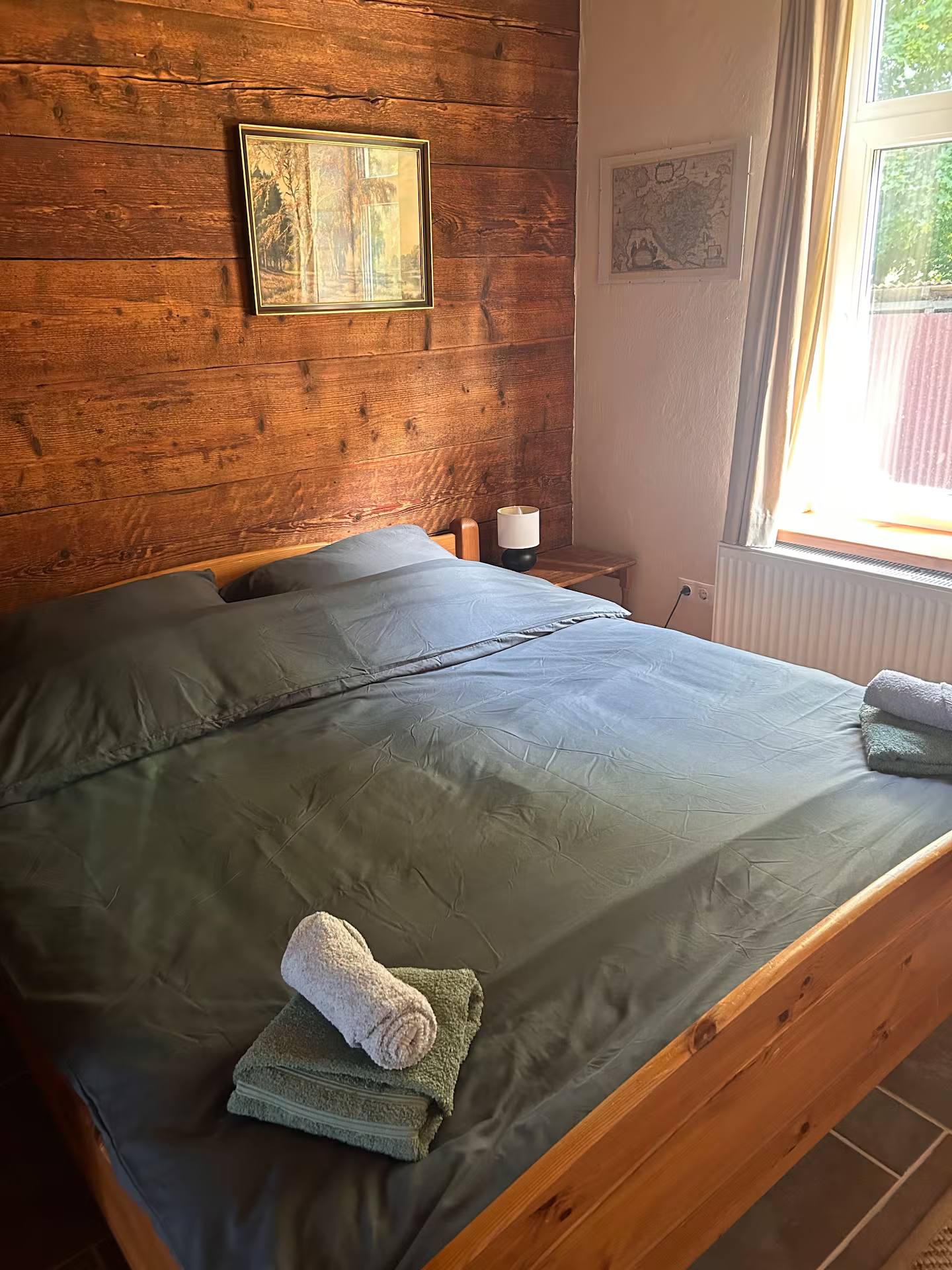 Doppelbett an der holzverschalten Wand, rechts ein Fenster mit Vorhang und ein Heizkörper; über dem Bett zwei gerahmte Bilder.