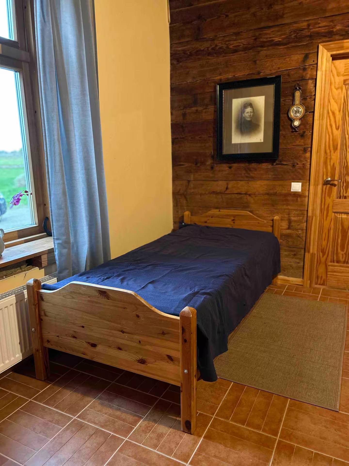 Einzelbett mit blauer Tagesdecke vor holzverschalter Wand, daneben eine Holztür und ein gerahmtes Bild, links ein Fenster mit Heizkörper.