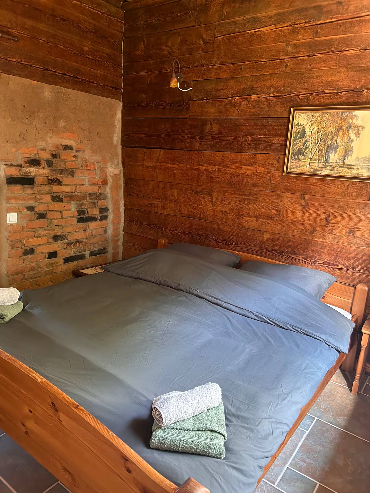 Schlafraum mit Doppelbett aus Holz vor einer holzverschalten Wand mit offenem Ziegelfeld; auf dem Bett liegen gefaltete Handtücher, der Boden hat Terrakottafliesen.