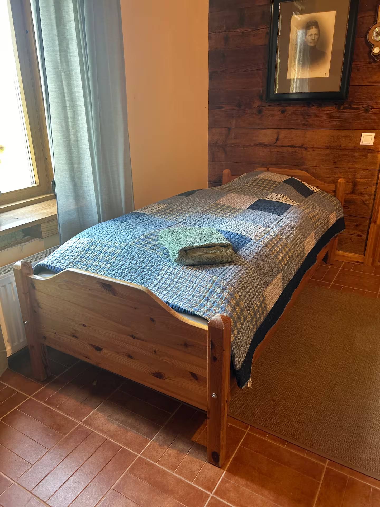 Einzelbett aus Holz mit blau gemusterter Tagesdecke an einer holzverschalten Wand, links ein Fenster mit grünem Vorhang.