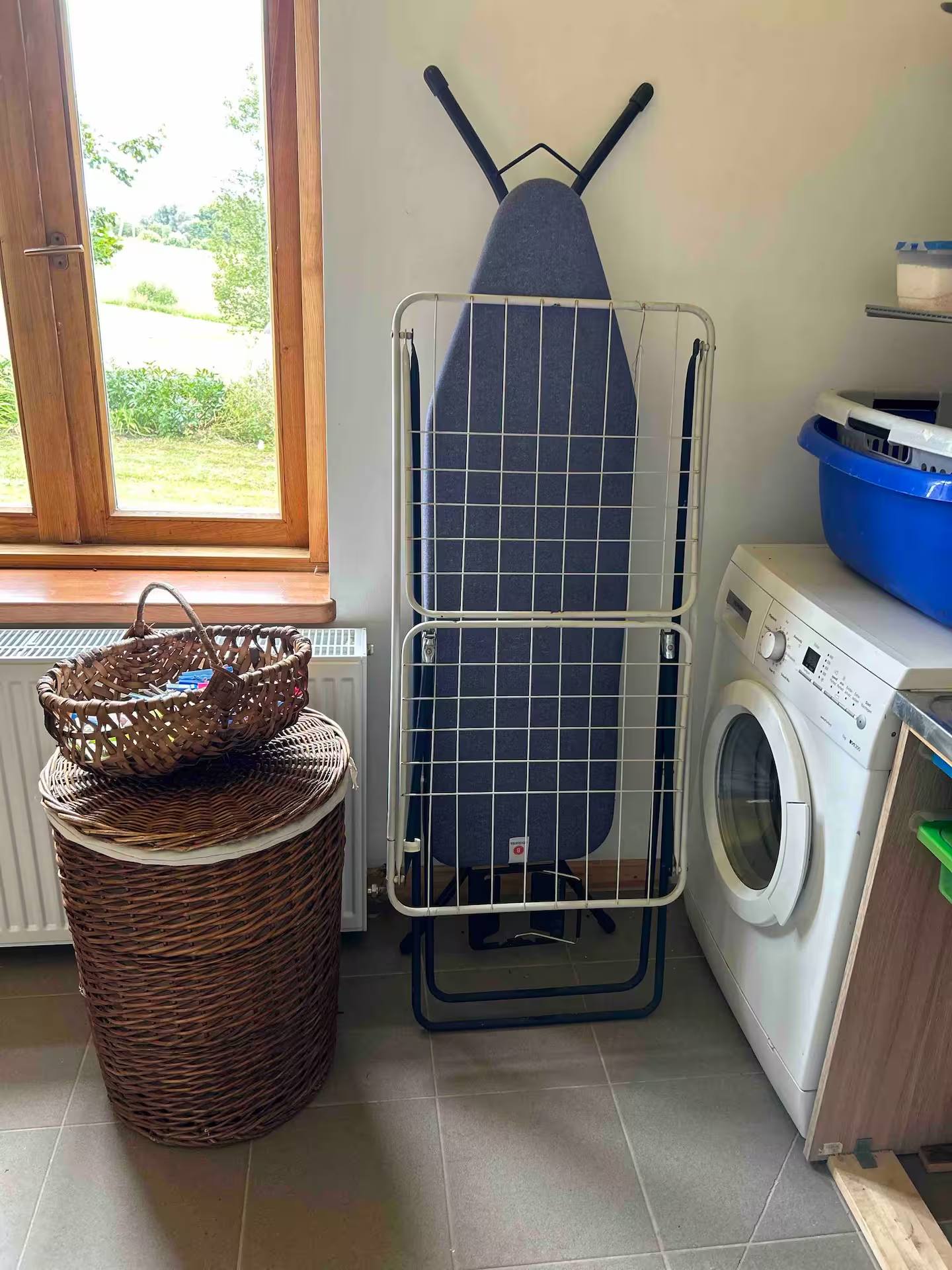 Waschmaschine unter einer Arbeitsplatte, davor ein aufgestelltes Bügelbrett, ein Wäschekorb aus Weide und Waschmittelbehälter.