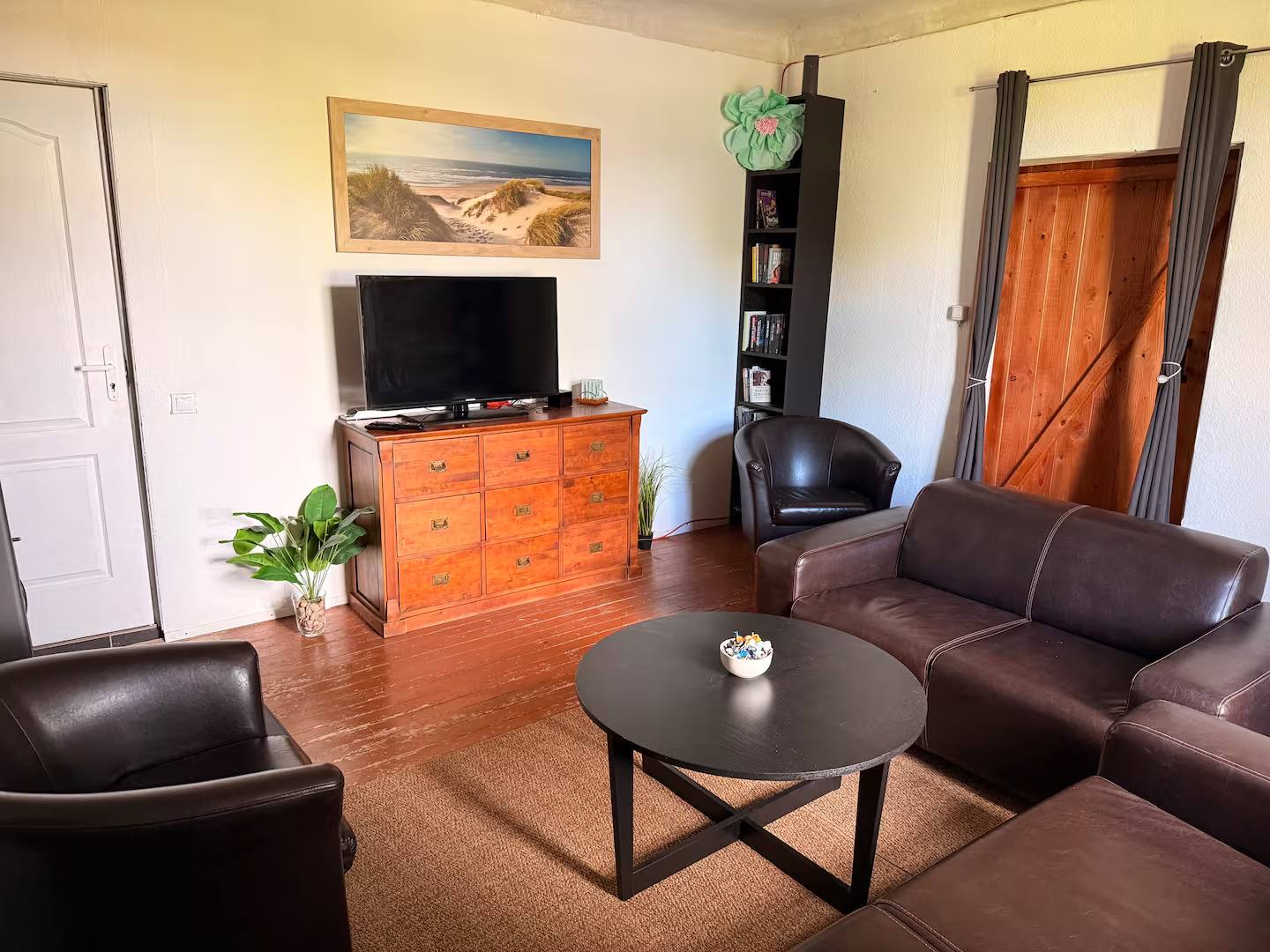 Zweiter Blick in das Wohnzimmer: Fernseher auf einer Holzkommode, Ledersofa und Sessel um den Couchtisch, Regal und Holztür.