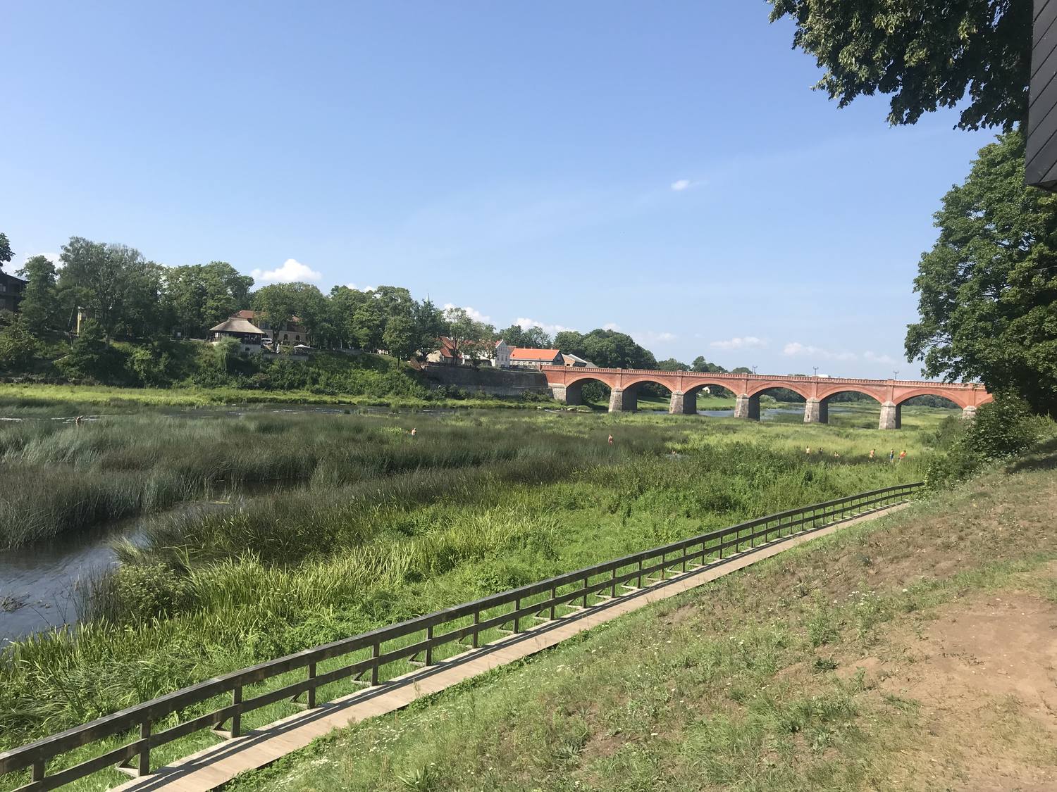 Mehrbogige Brücke aus roten Ziegeln über einen Fluss, im Vordergrund ein Holzsteg und Schilf, links eine Wiese.
